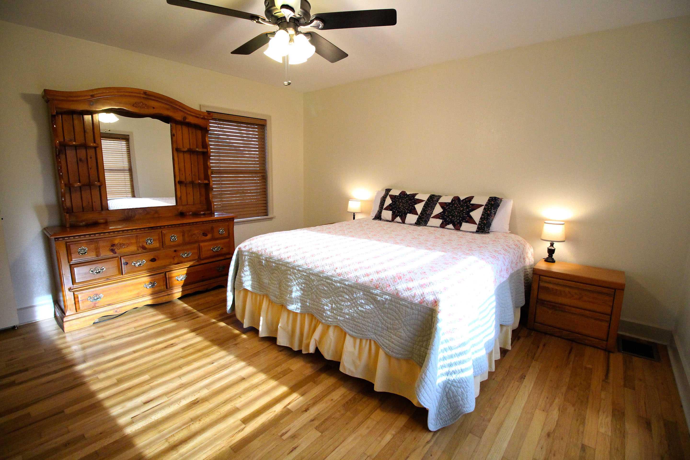 A wide view of a guest room with a bed, dresser, nightstand, and ceiling fan.