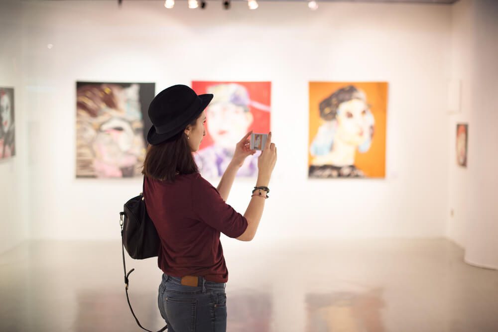 A woman stands in a gallery, taking a photo, at First Friday Art Walk.